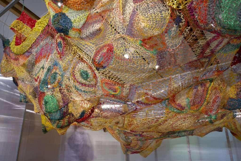 Una fotografia di un’opera realizzata con sottili corde di vari colori, sospese dal soffitto come una rete da pesca.