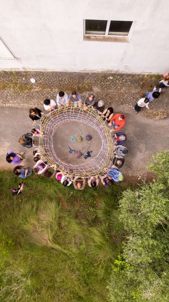 Fotografia dall’alto di un gruppo di persone che partecipano a un’attività artistica: ciascuno tiene in mano una parte di una rete colorata, formando un cerchio.